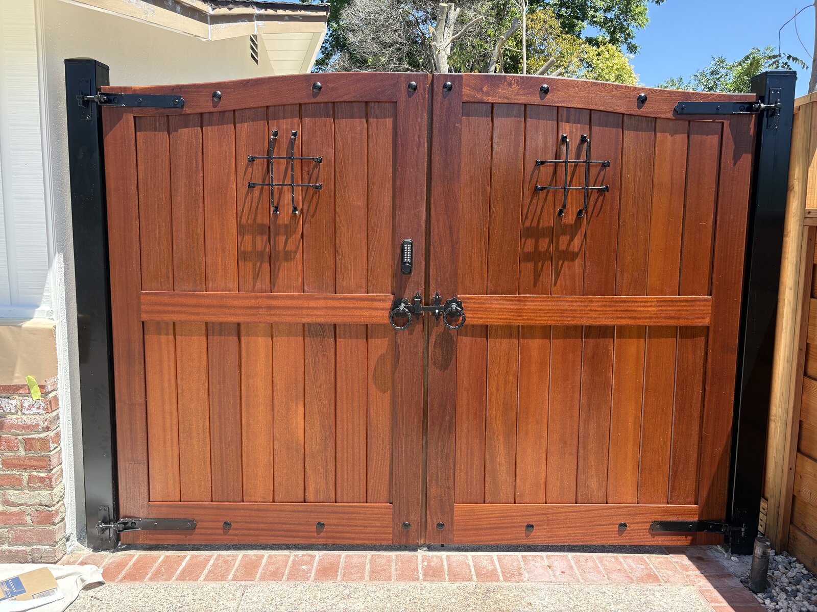 Double mahogany driveway gate with iron speakeasy grilles, ring pulls, keypad lock and black steel frame