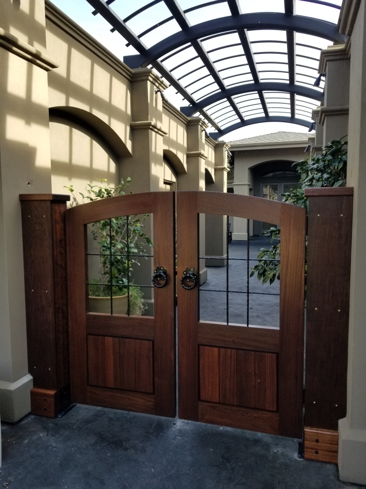 Double swing mahogany entryway gate with decorative iron inlay grille, ring pulls, and arched tops under a curved glass canopy
